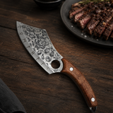 A handmade cleaver-style knife with a patterned steel blade and a brown wooden handle, displayed diagonally on a dark wooden table with a plate of sliced steak in the background.
